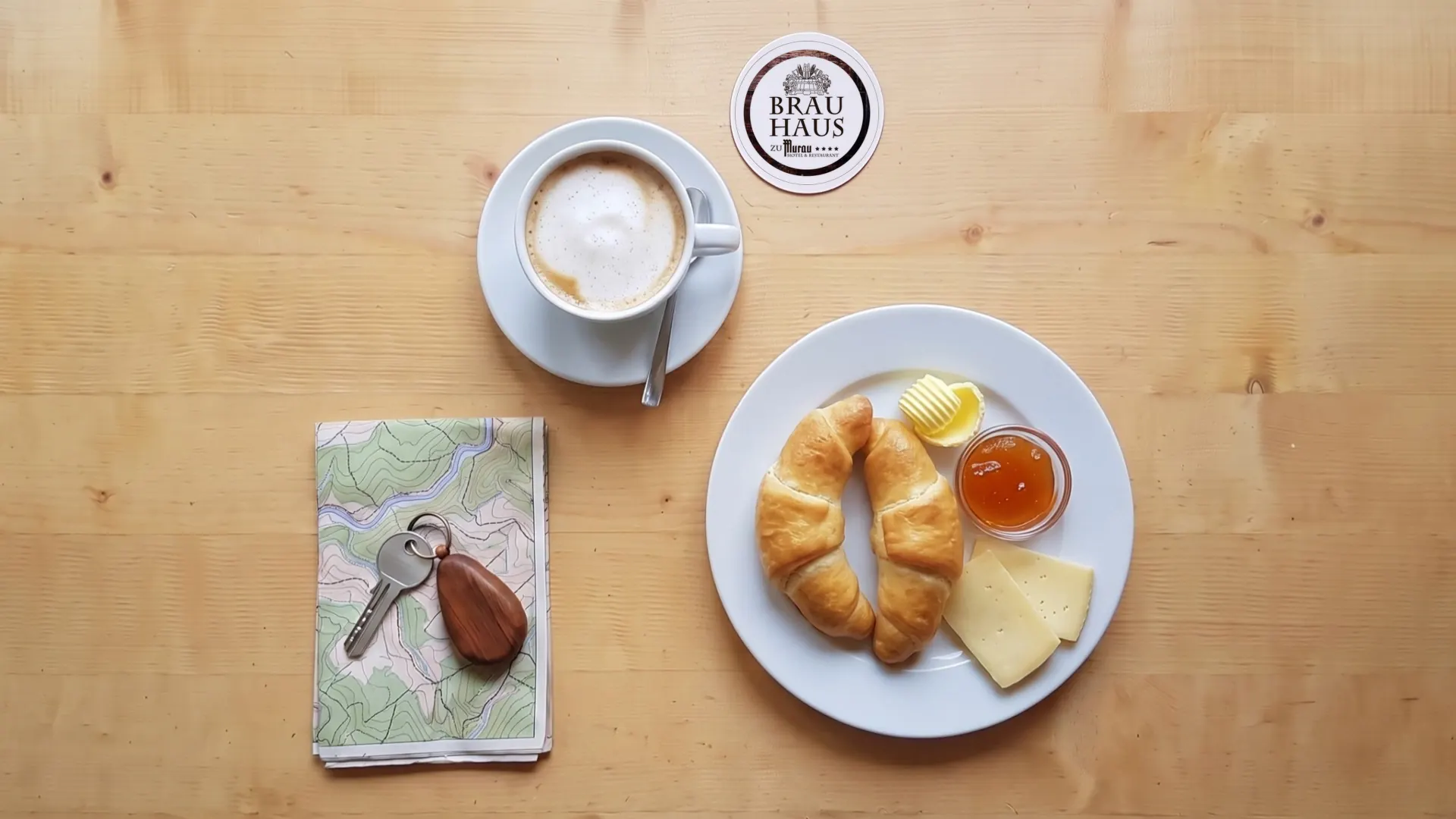 Derselbe Tisch am nächsten Morgen im Tageslicht: Kaffee, Kipferl und Frühstücksteller, daneben wieder der Bierdeckel mit dem Brauhaus-Logo.